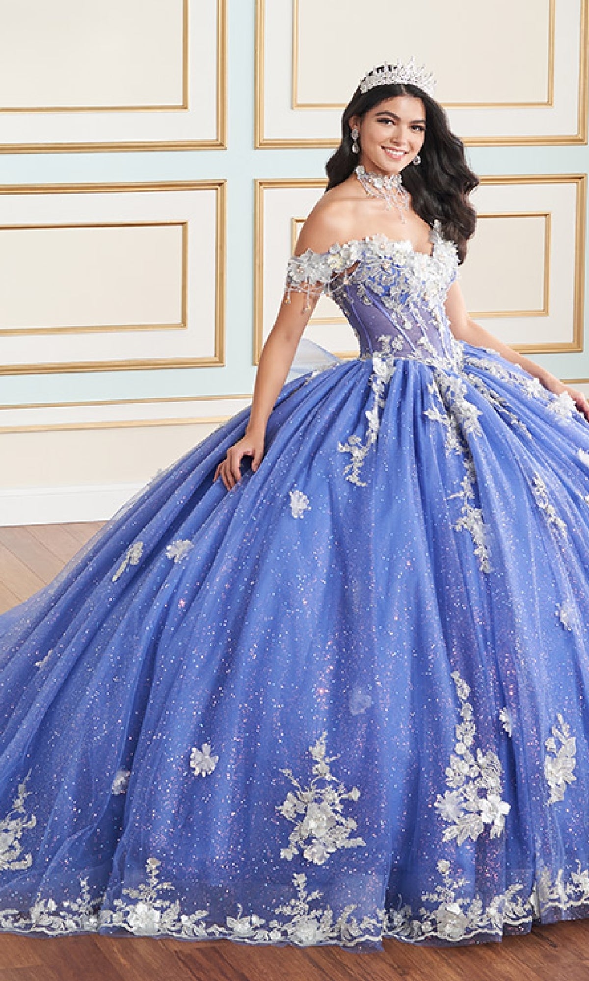 A young woman in the Quinceanera Dress PR30178 By Princesa, a sparkling blue gown with white floral details and a corset bodice, wearing a tiara, stands in a formal room with decorative paneling.